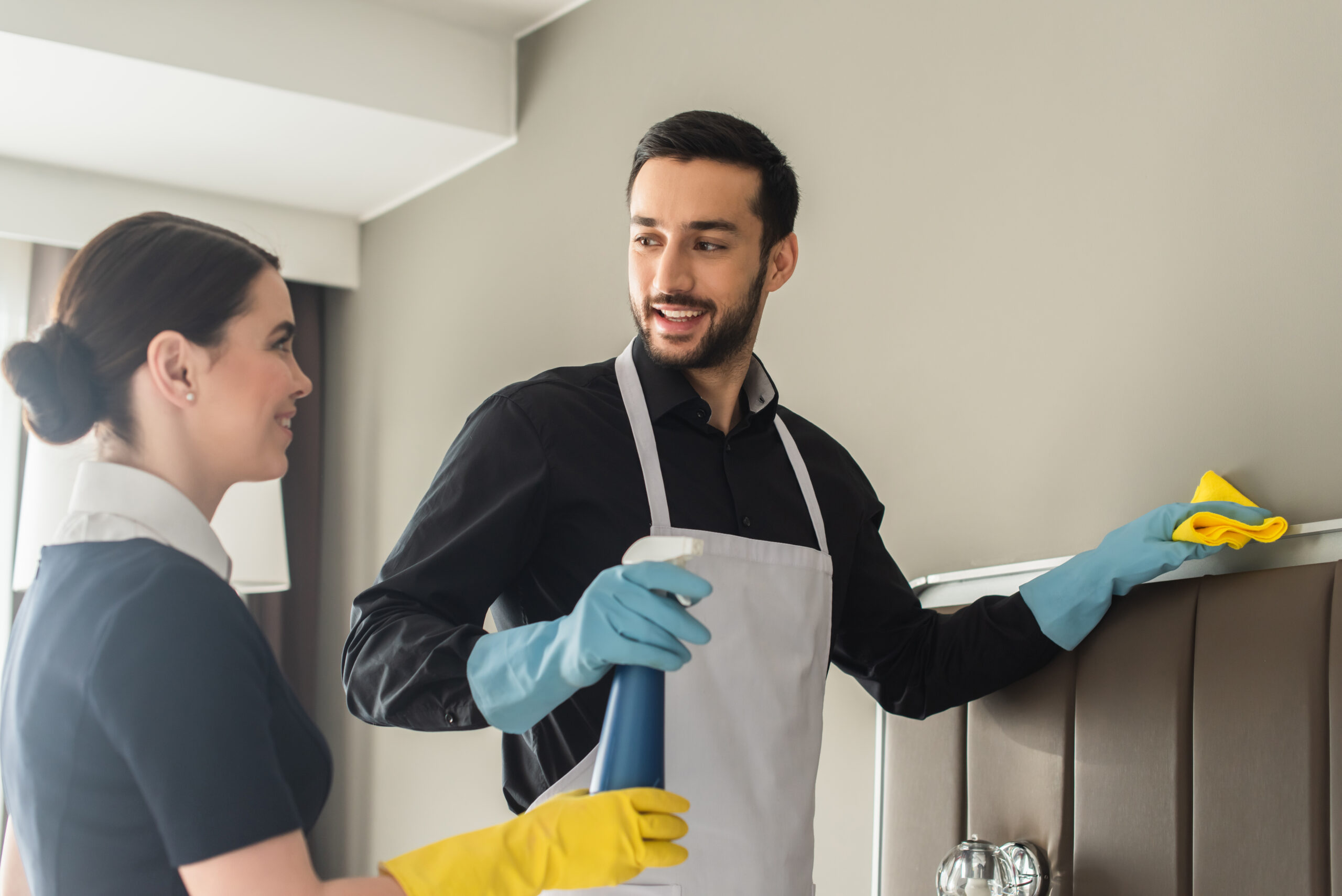 cheerful housekeepers looking at each other while holding cleaning supplies and cleaning hotel room