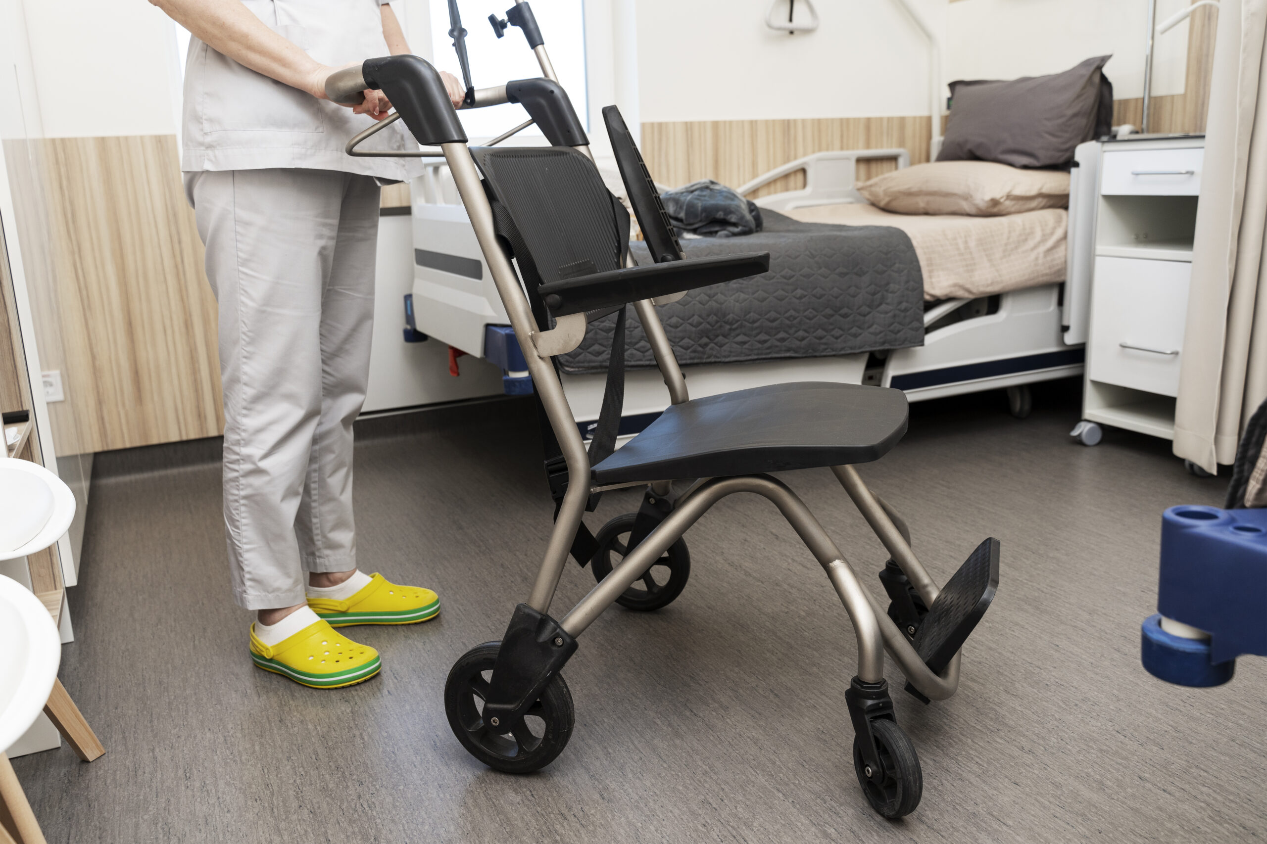 Helping a patient get into bed in a hospital ward. A nurse holds a wheelchair by the handle.